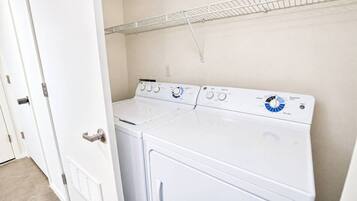 A laundry room with a front-loading washer and dryer, a metal shelf above, and a white door.
