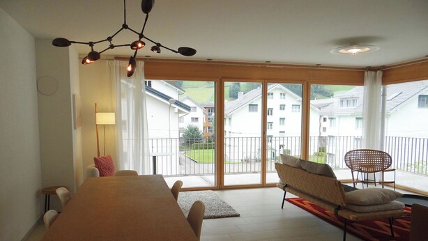 A spacious room with a large dining table, a sofa, and a chair. There is a balcony with a view of houses and greenery.