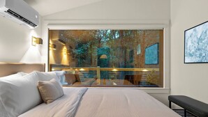 A modern bedroom with a large window, a bed with white linens, a bench, and a wall-mounted air conditioner. - Romantic Hideaway | Hot Tub Near Thunder Ridge (Omaha)