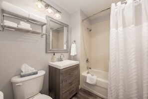 Un baño con inodoro, lavamanos, tocador, espejo, toallero, ducha y cortina.