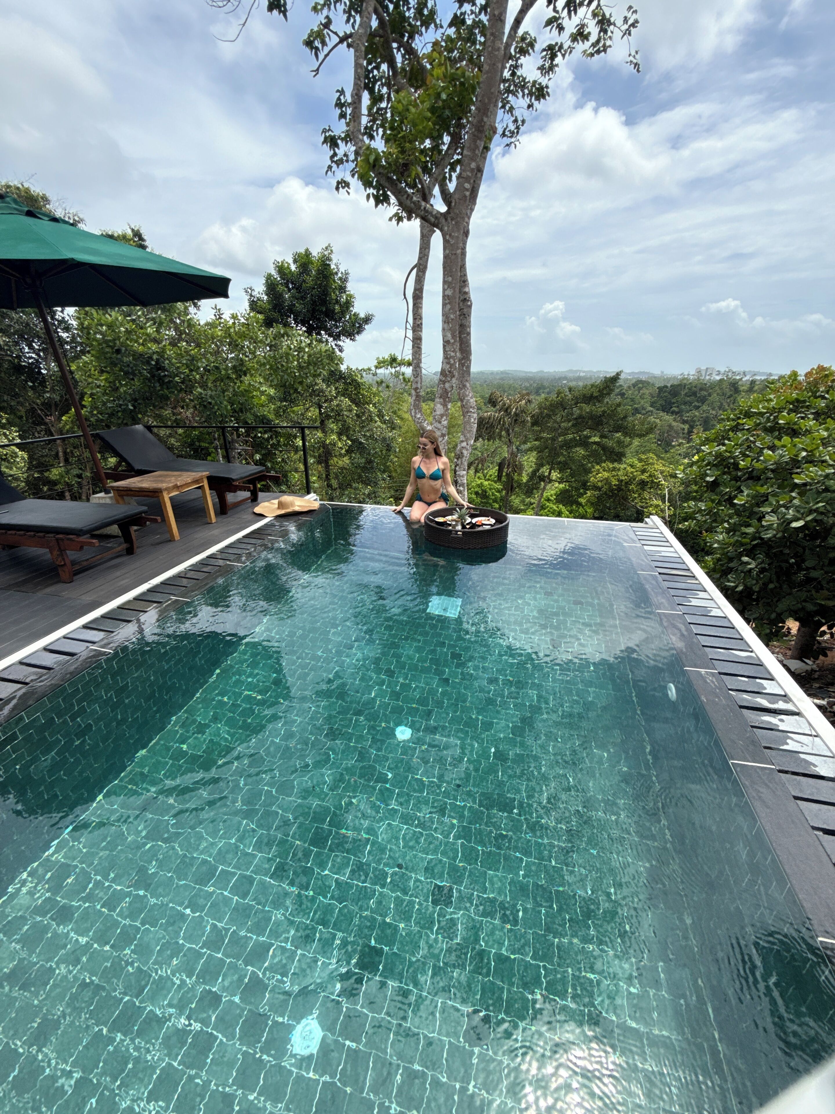 A poolside area with a person sitting by the pool, surrounded by lush greenery and a clear view of the landscape.