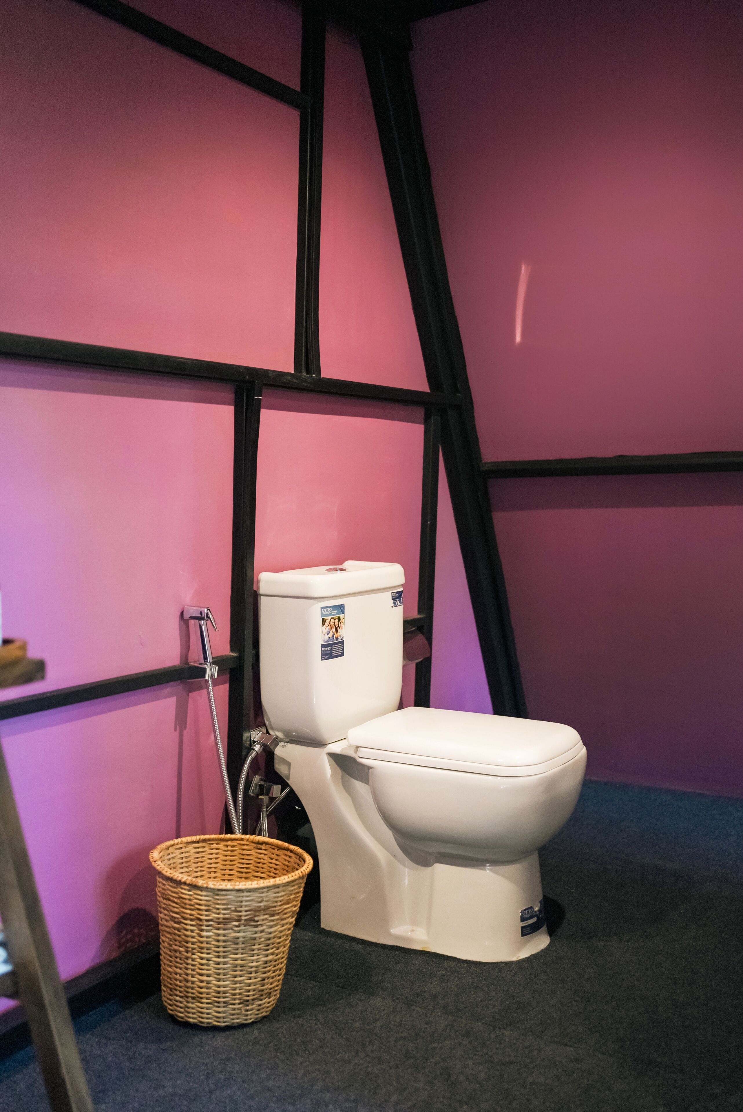 Et badeværelse med et hvidt toilet, en fletkurv og en lyserød væg med sort indramning.
