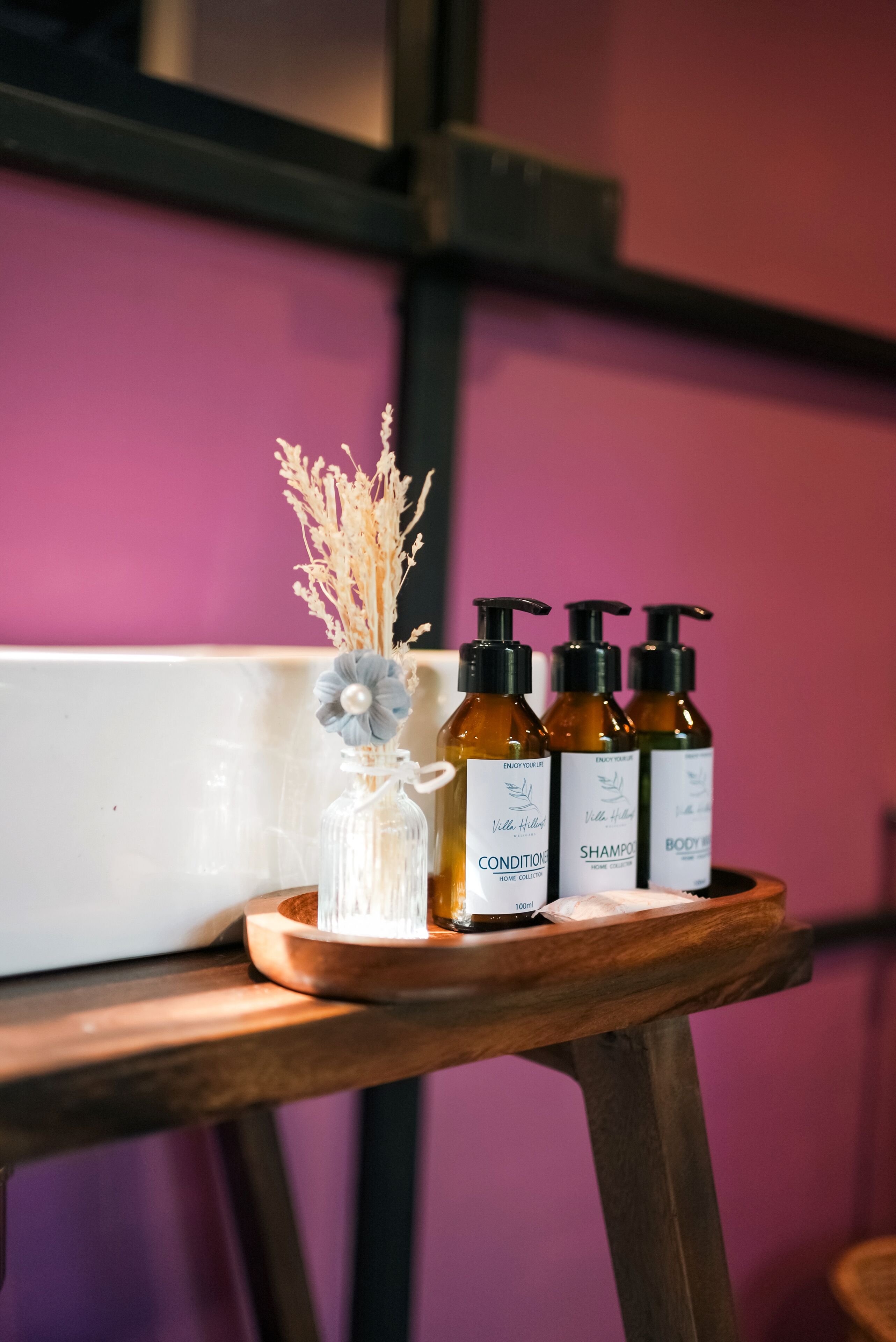 A wooden table with three bottles of body care products and a clear glass bottle with dried flowers.