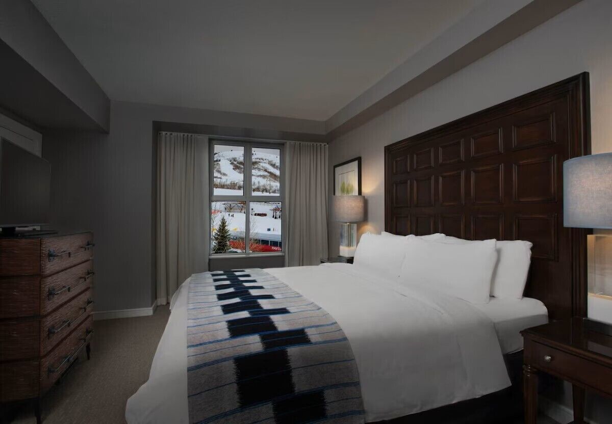 Habitación de hotel con una cama grande, cómoda, ventana con cortinas y cabecera de madera.