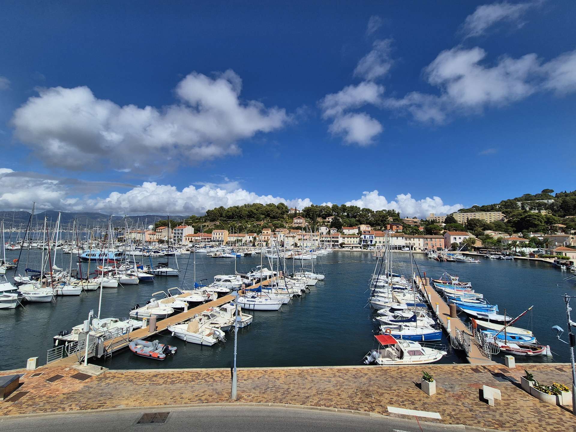 Un port de plaisance où sont amarrés de nombreux bateaux, entouré de bâtiments et d’un paysage vallonné.