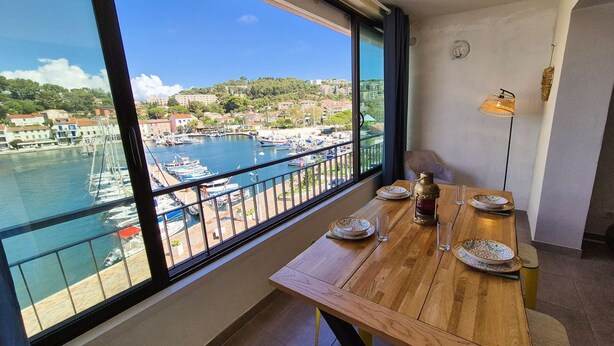 A balcony with a wooden table set for dining, overlooking a marina with boats and buildings. - 2-Bedroom Apartment with Sea View – Everything Within Walking Distance (ST MANDRIER SUR MER)
