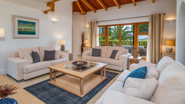Amplia sala de estar con dos sofás, una mesa de centro y vistas a un paisaje tropical a través de amplios ventanales.