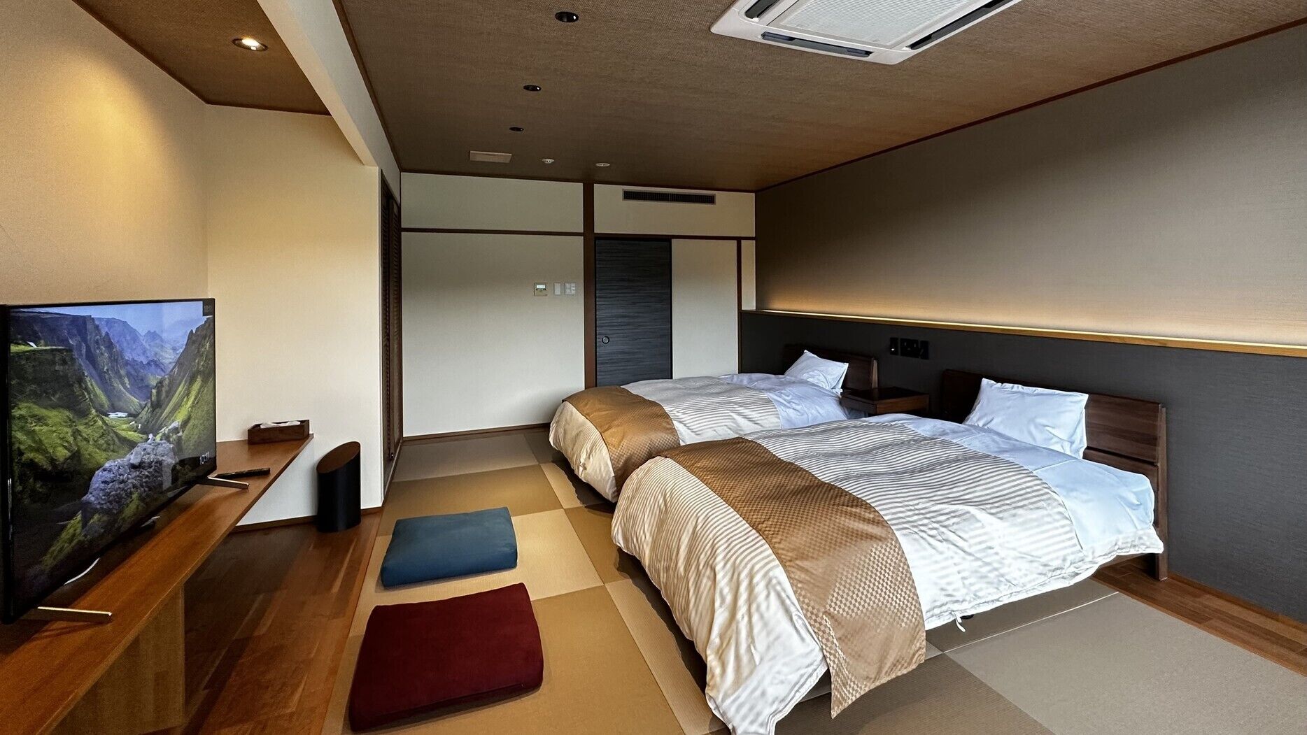 A modern hotel room with a large flat-screen TV, two beds with matching headboards, a wooden desk, and a large window.