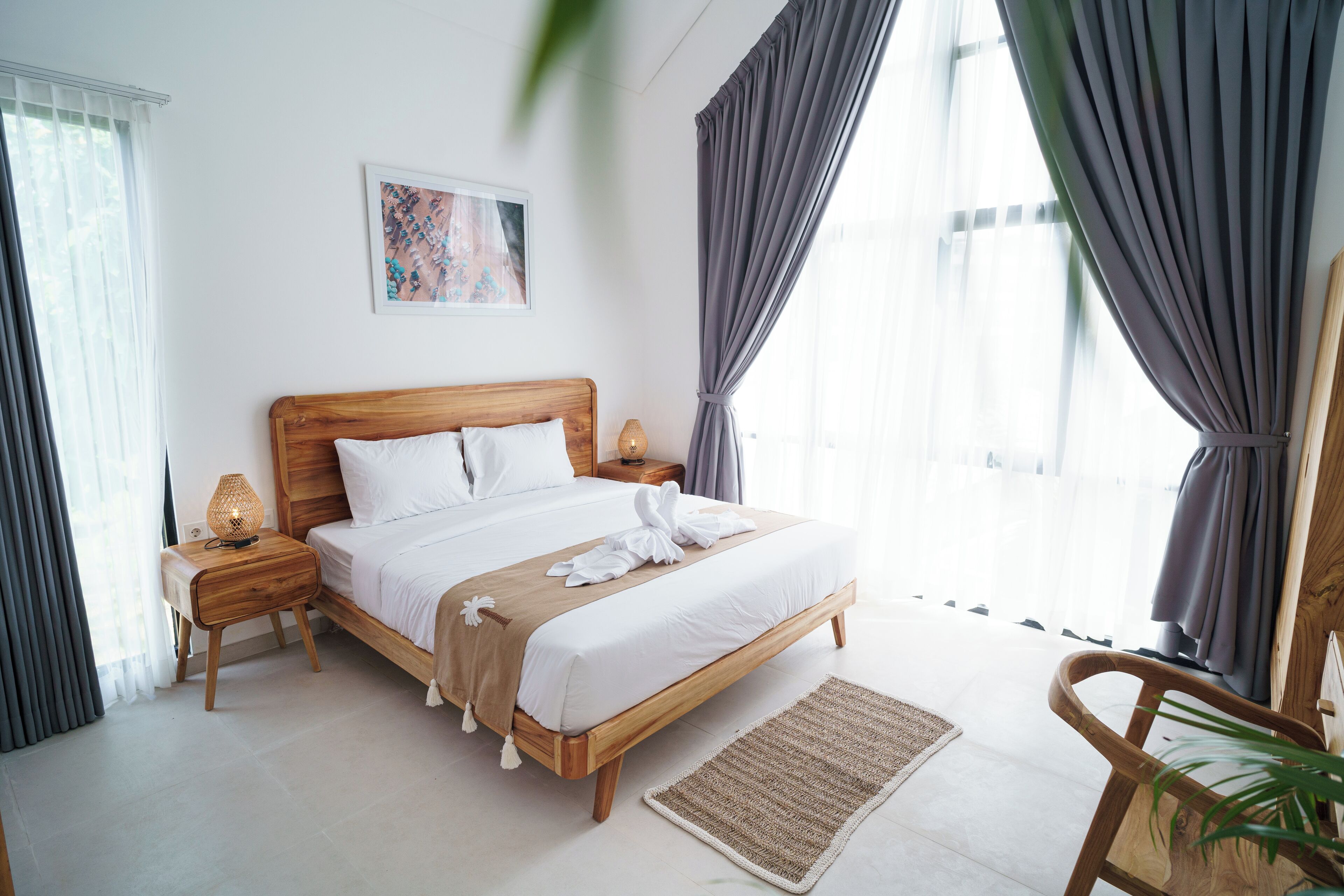 A neatly made bed with a wooden headboard, a bedside lamp, and a small table with a lamp. There is a framed picture on the wall and a chair with a plant.