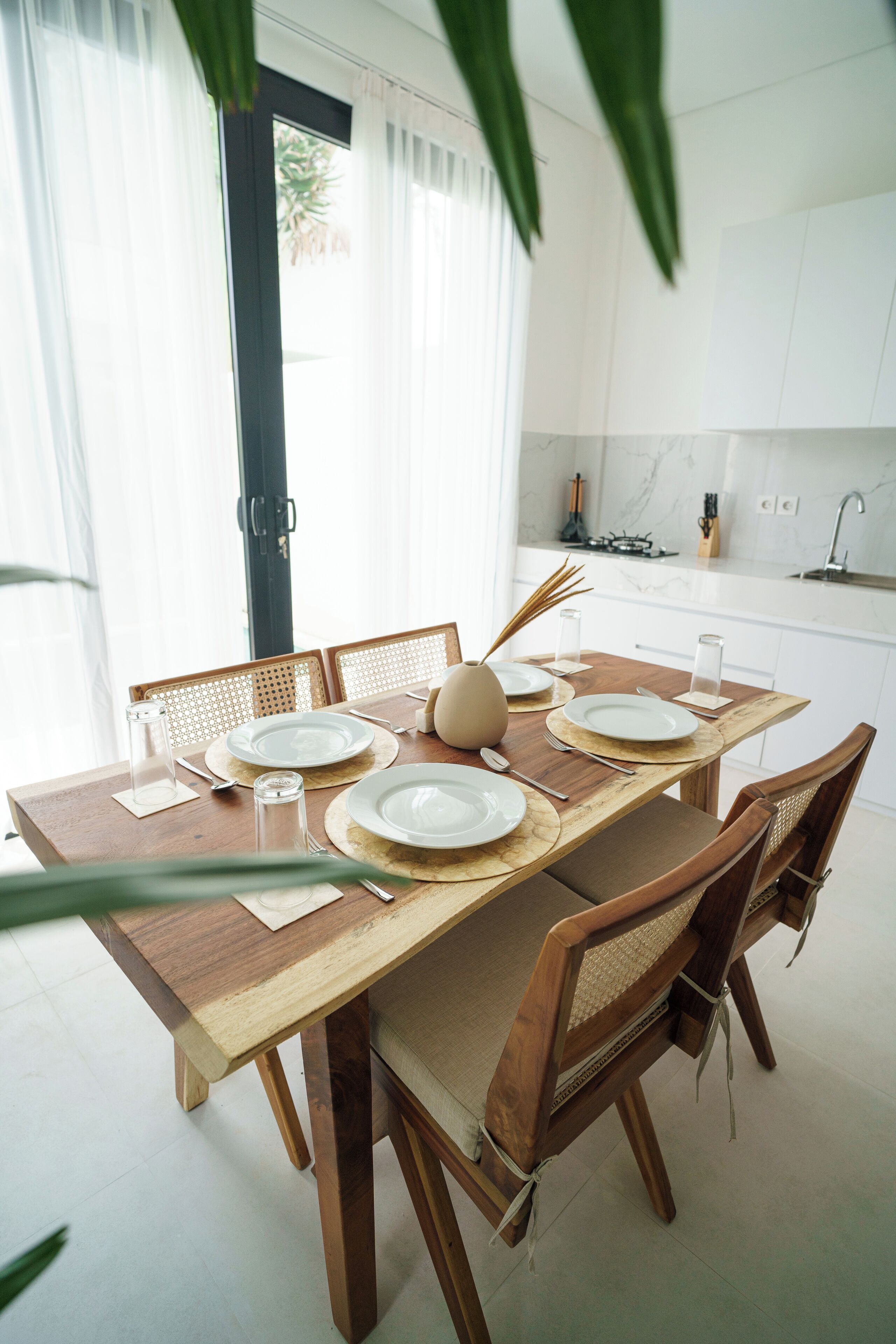A dining table set with plates, glasses, and cutlery in a modern kitchen with white cabinets and a large window.