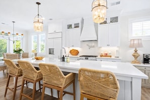 Une cuisine moderne avec des meubles blancs, un grand îlot central doté de tabourets, des luminaires suspendus et une vue sur la verdure à l’extérieur.