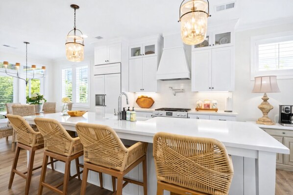 A modern kitchen with white cabinetry, a large island with bar stools, pendant lighting, and a view of greenery outside.