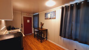 A compact kitchen with a gas stove, sink, and a small table with stools. There is a red door and a painting on the wall. - SHERWOOD ARMS MOTEL_CABIN #2 (Running Springs)