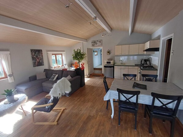 Un amplio espacio habitable con sofá, mesa de comedor y sillas, una cocina con armarios y electrodomésticos, y un techo con vigas a la vista.