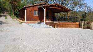 A small wooden cabin with a porch, gravel driveway, and surrounding trees. - Renovated chalet located by the Ourthe. (Tohogne)