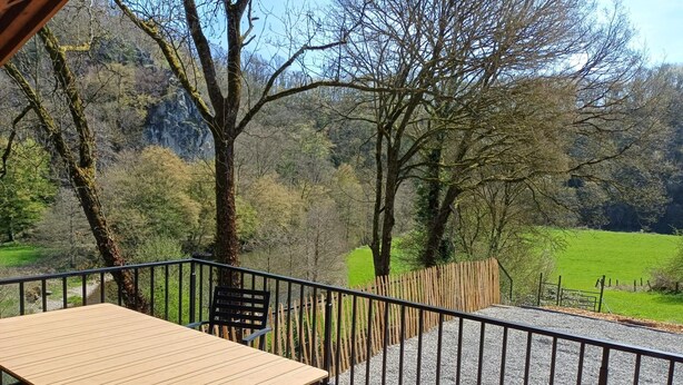 A wooden deck with a table and chairs, surrounded by a metal railing, overlooking a green landscape with trees and a distant hill. - Renovated chalet located by the Ourthe. (Tohogne)