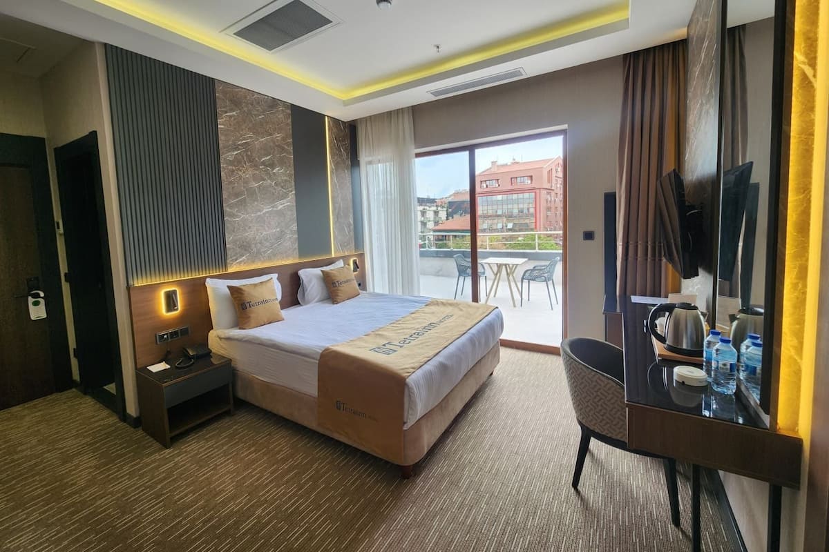 A hotel room with a bed, bedside table, desk, chair, and a view of the city through the window.