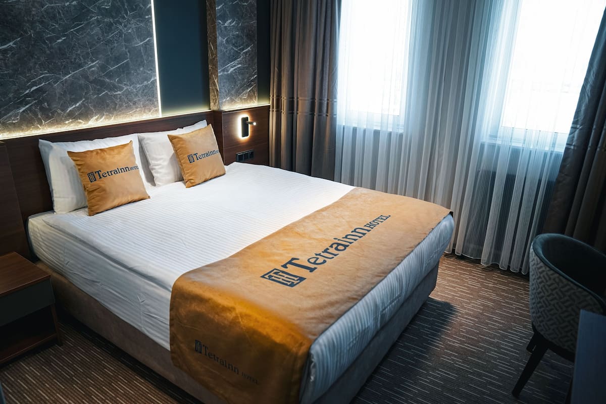 A hotel room with a large bed, two pillows with the hotel logo, a nightstand, a chair, and a window with curtains.