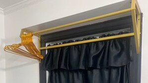 Ein Zimmer mit einem Fenster, das mit schwarzen Vorhängen verdeckt ist, einem weißen Schreibtisch mit einem türkisblauen Handtuch und einem goldfarbenen Wäscheständer.