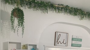 Ein Schlafzimmer mit einem weißen Bett, grün-weißer Bettwäsche, einem Schreibtisch mit Lampe und einer hängenden Pflanze.