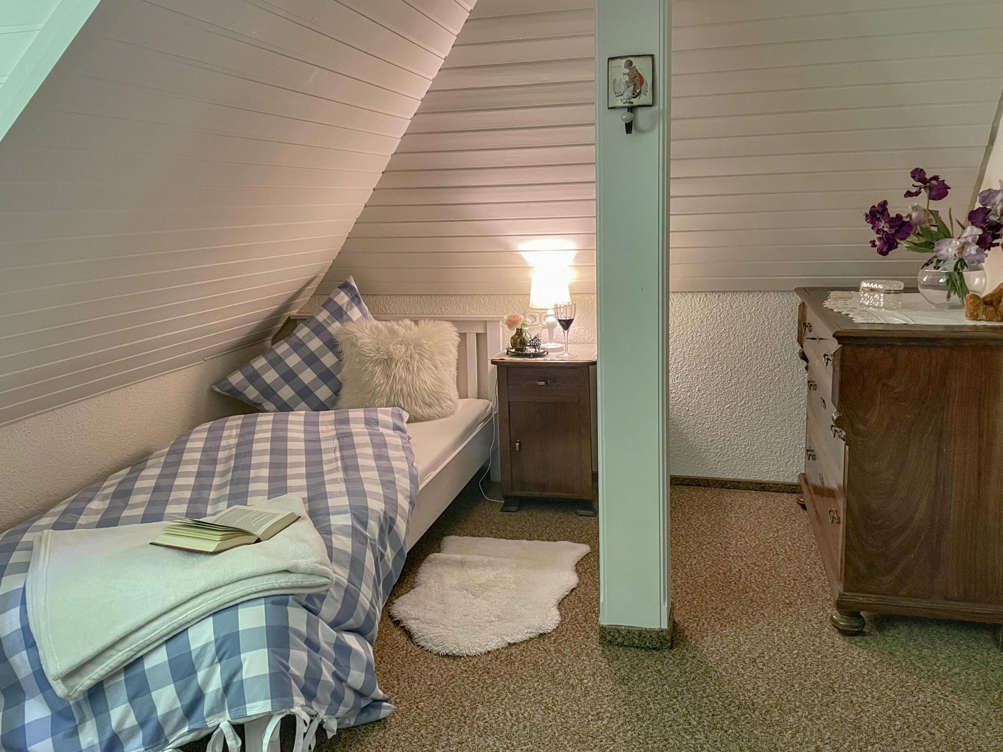 Une chambre avec un plafond en pente, un lit avec une literie à carreaux, une table de chevet avec une lampe, une commode et une petite table avec des fleurs.