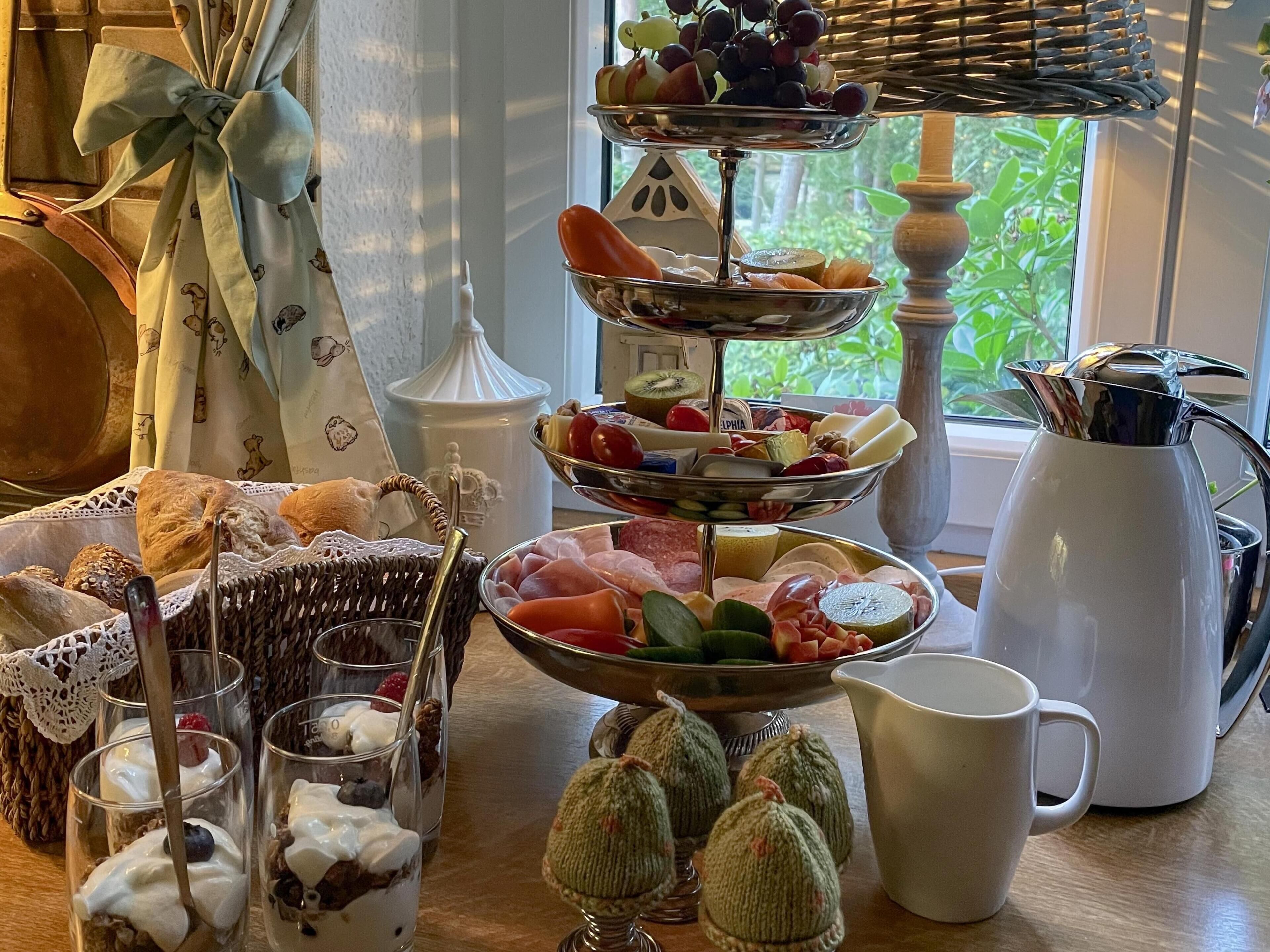 Un buffet della colazione con diversi cibi, un cesto di vimini, un boccale e una finestra con piante verdi all'esterno.