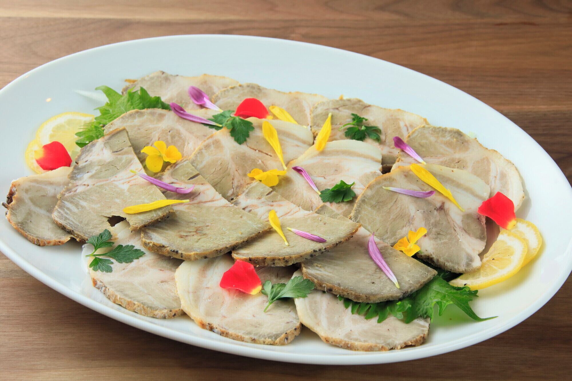 Un piatto bianco con fette di carne guarnito con prezzemolo, fette di limone e fiori eduli.