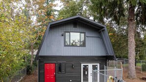 Una cabaña pequeña y oscura de color oscuro, con una puerta roja y una ventana, rodeada de árboles y una zona cercada.