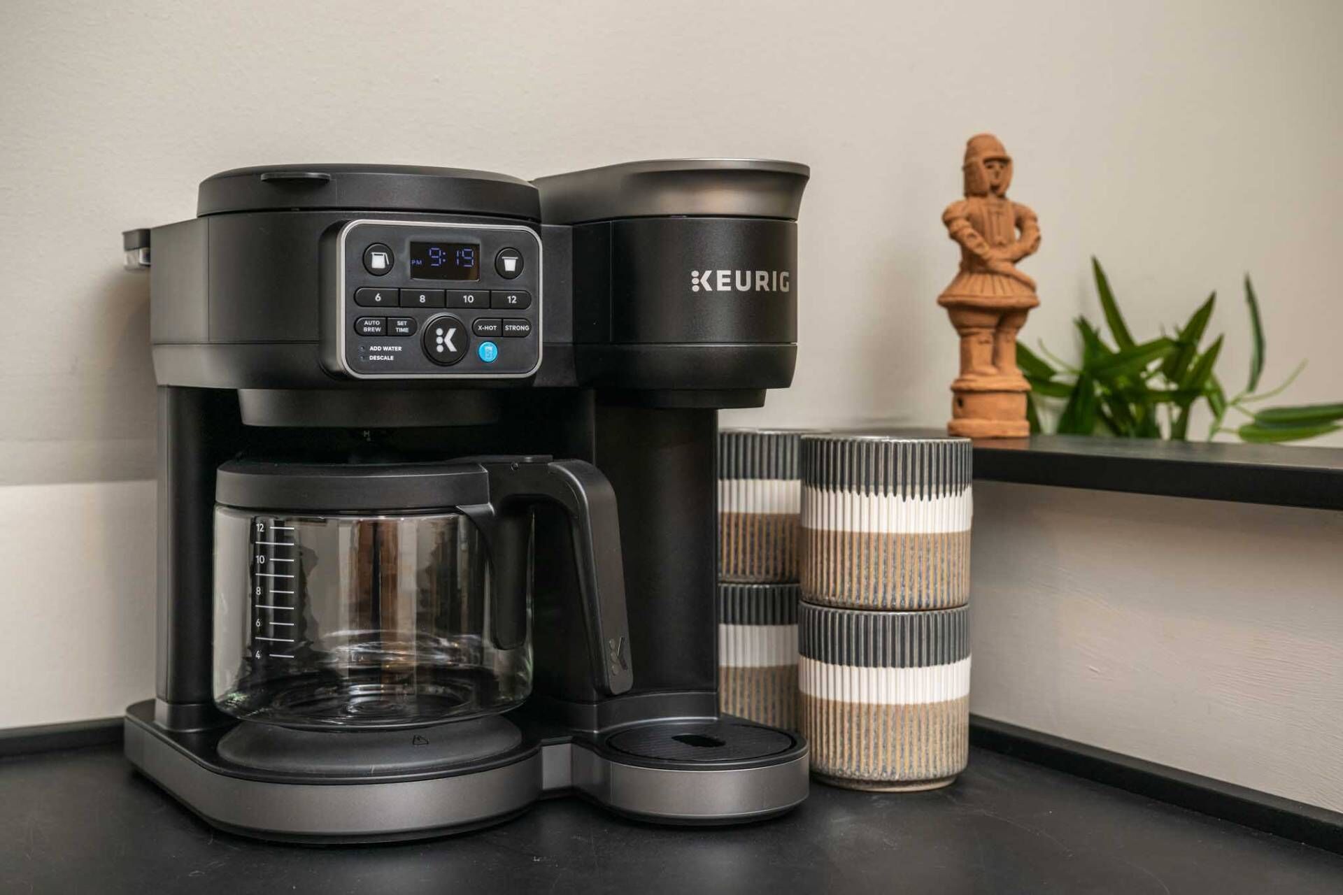 Una cafetera Keurig con pantalla digital, una jarra y dos vasos de viaje apilados sobre una encimera.