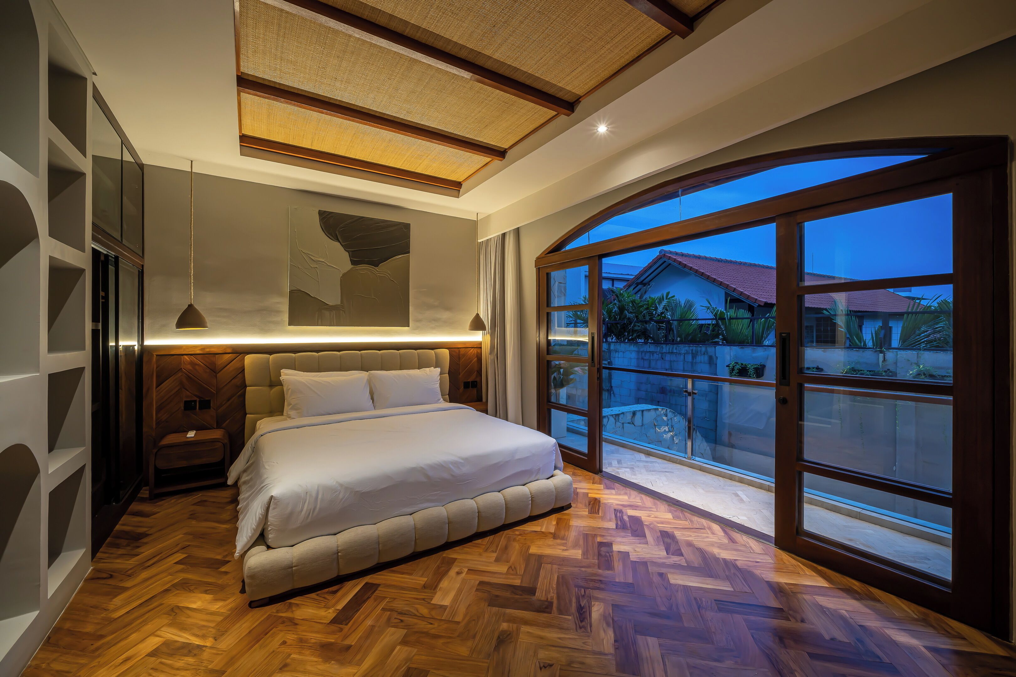 A modern bedroom with a large bed, wooden flooring, and a view of a residential area through large windows.
