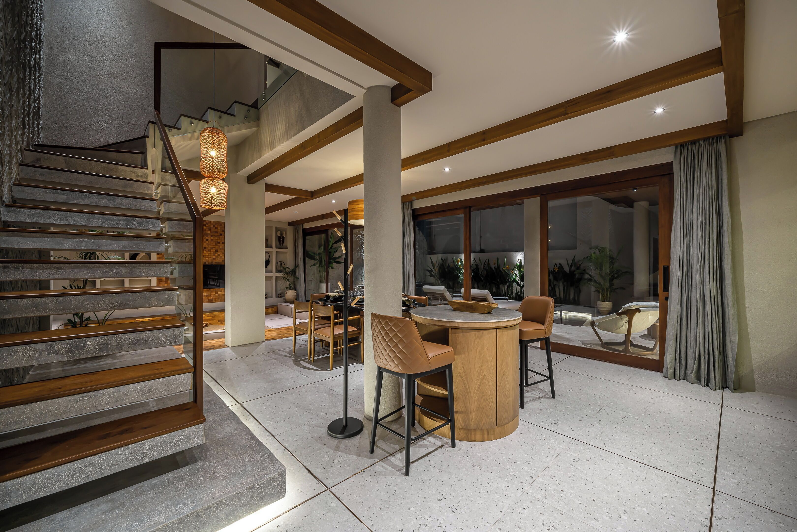 Un interior moderno con una escalera, vigas de madera, una mesa redonda de madera con sillas y un amplio ventanal con cortinas.