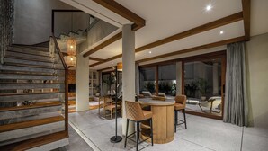 Un interno moderno con una scala, travi in legno, un tavolo rotondo in legno con sedie e un'ampia finestra con tende.