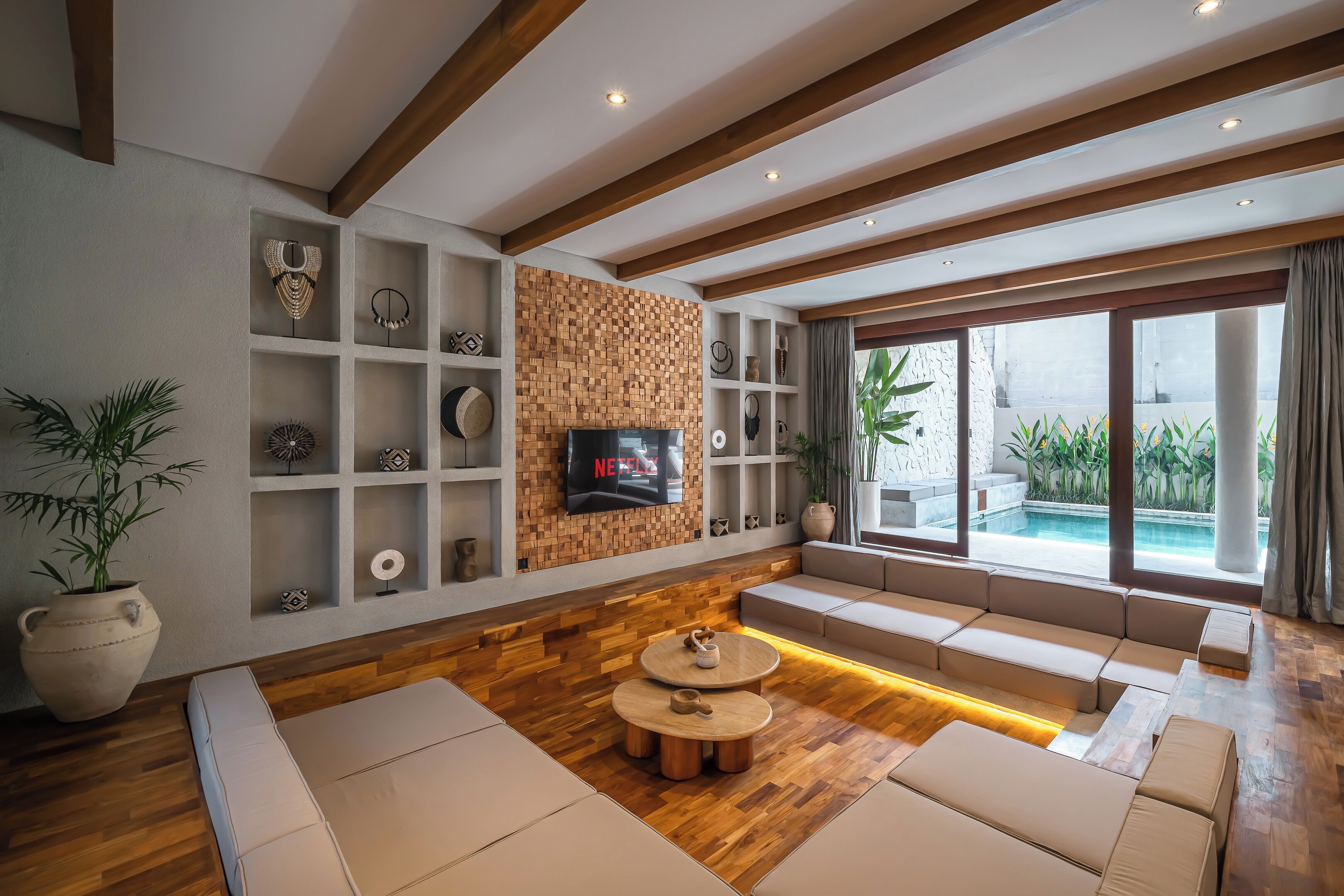 Una sala de estar moderna con piso de madera, estanterías empotradas, televisor y una zona de estar con vista a una piscina.