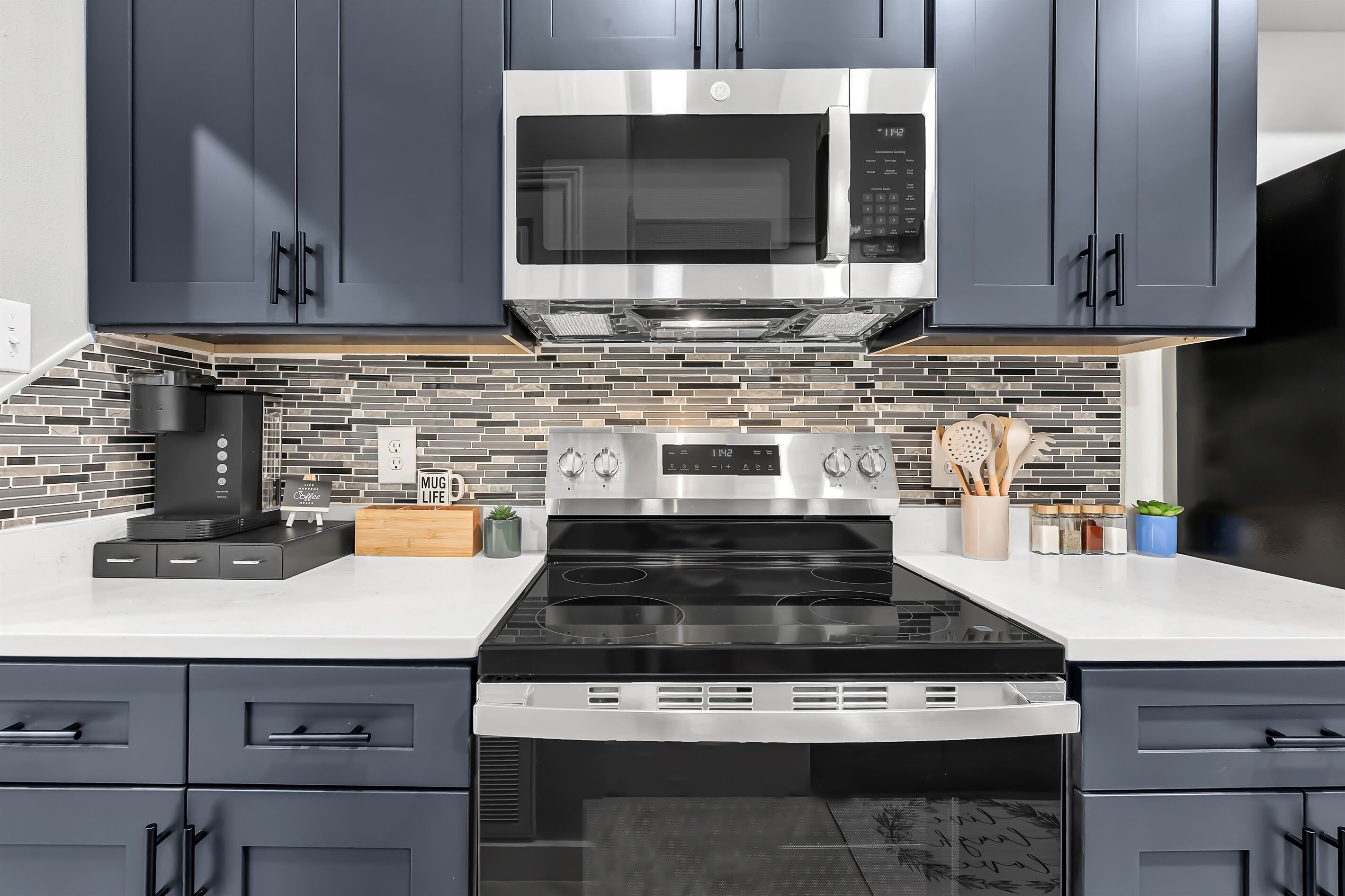 A modern kitchen with dark cabinets, a stainless steel microwave, a black stove, and a tiled backsplash.