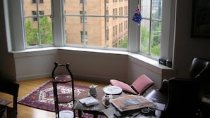 A room with a large bay window overlooking a cityscape, a small table with a teapot, cups, and a book, and a chair with a cushion.