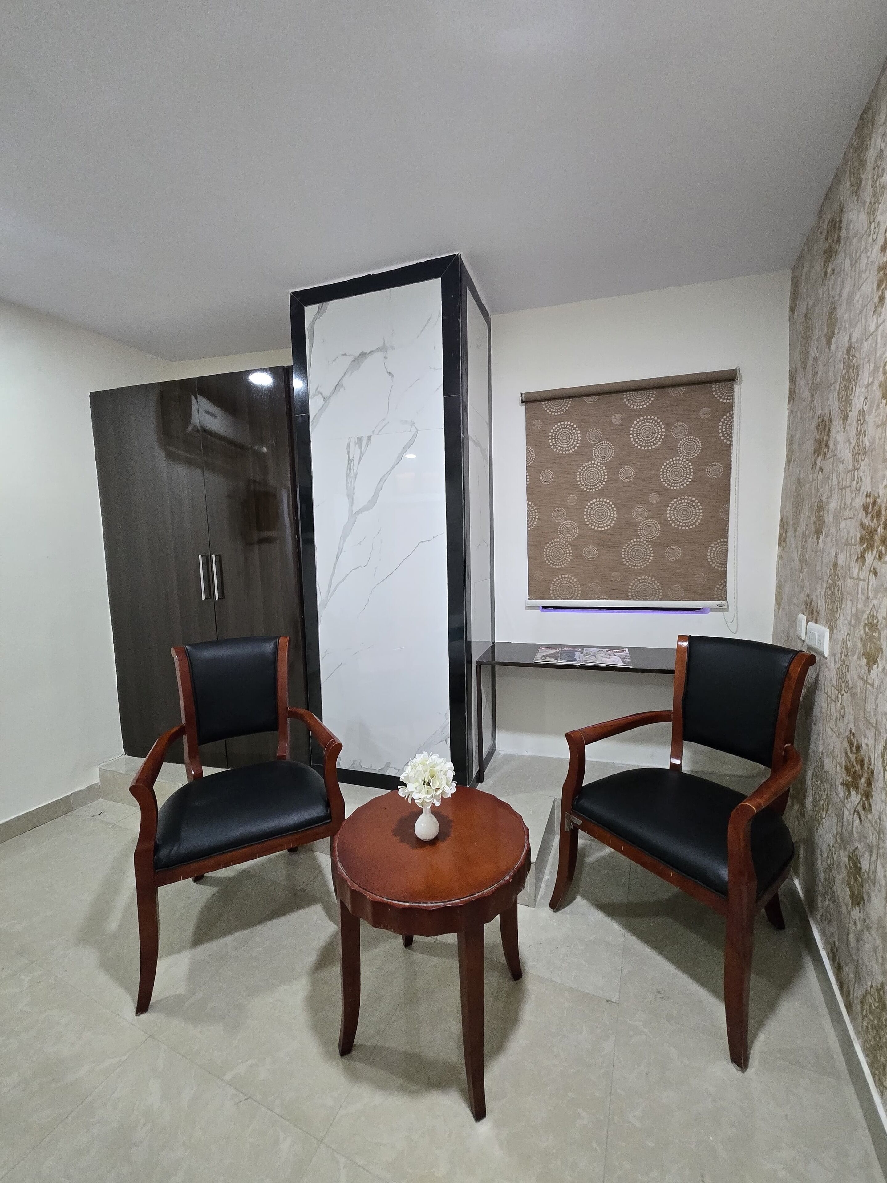 A room with two wooden chairs, a small round table with a vase, a cabinet, a television, and a patterned curtain.