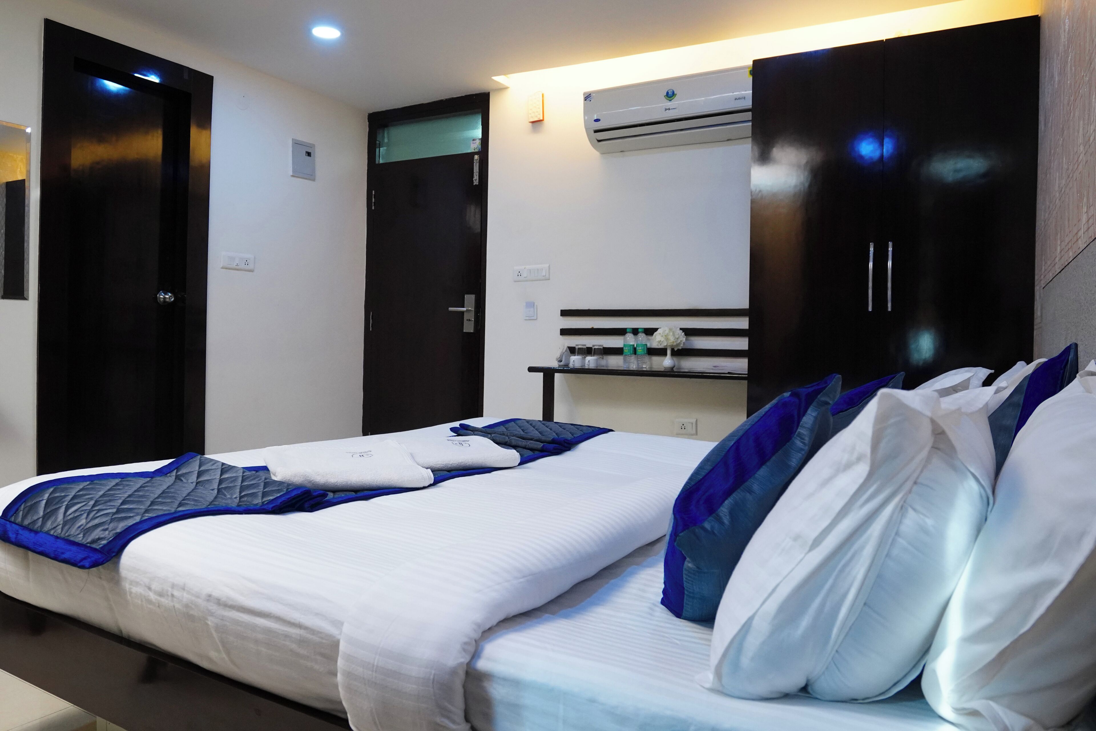 A hotel room with two beds, a dark wooden wardrobe, an air conditioning unit, and a small shelf with toiletries.