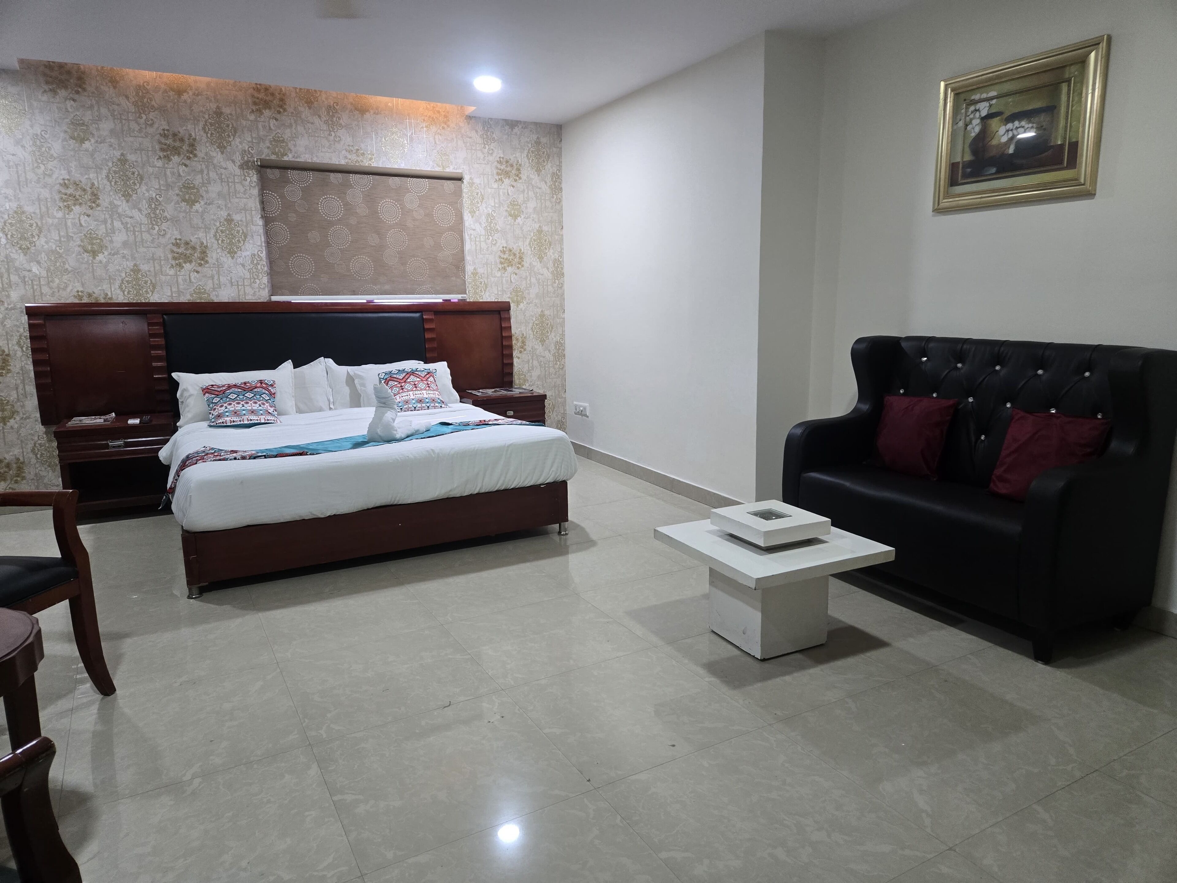 A hotel room with a bed, bedside tables, a black leather sofa, a small white table, and a framed picture on the wall.