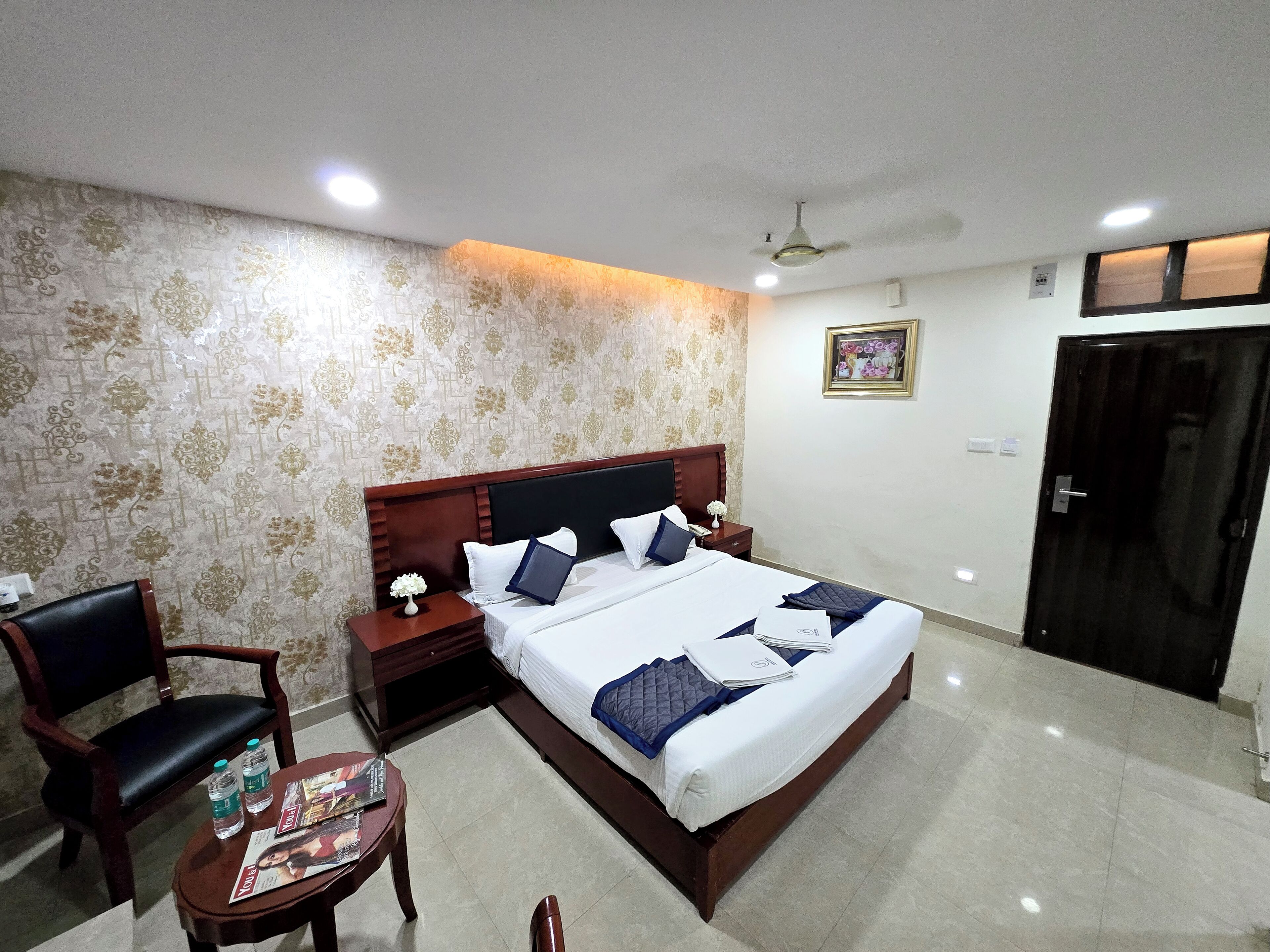 A neatly arranged hotel room with a large bed, a wooden nightstand, a chair, and a small table with magazines and water bottles.