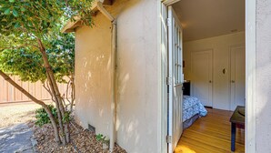 A room with a bed, a wooden floor, and a door leading to an outdoor area with a tree and a fence. - Serene Studio in SF Suburbs: Quiet + Cat Friendly! (Castro Valley)