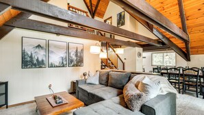 A spacious living area with a high ceiling, wooden beams, a large sectional sofa, a dining table with chairs, and framed artwork on the walls. - Sleek Blue Lake Springs Retreat w/ Deck & Yard! (Arnold)
