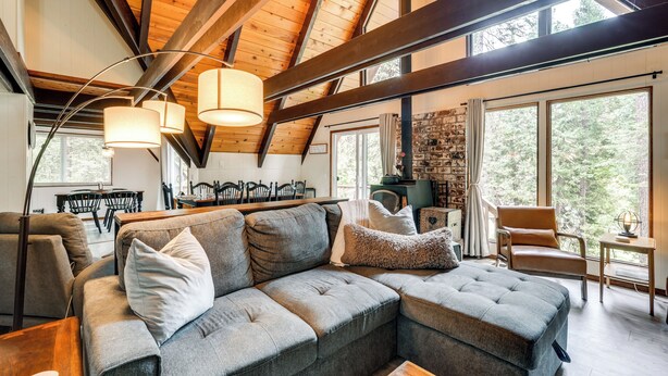 A cozy living room with a sectional sofa, wooden beams, large windows, and a stone fireplace. - Sleek Blue Lake Springs Retreat w/ Deck & Yard! (Arnold)
