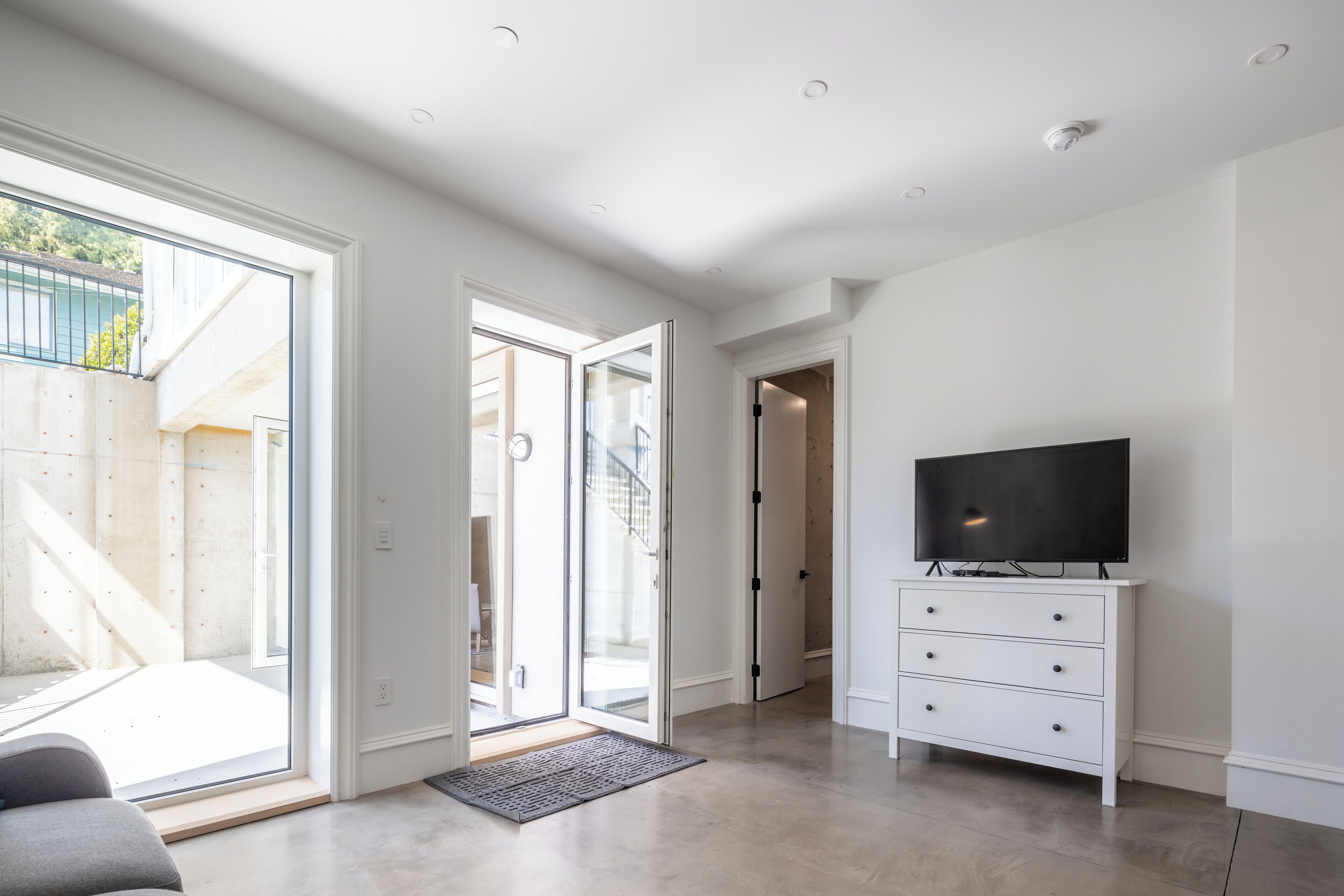 Un salon moderne doté d’une grande porte-fenêtre coulissante donnant sur un balcon, d’un téléviseur à écran plat posé sur une commode blanche et d’un escalier visible à travers la porte.
