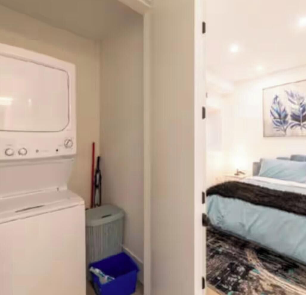 A compact room with a washing machine, a small trash bin, and a bed with a patterned bedspread.