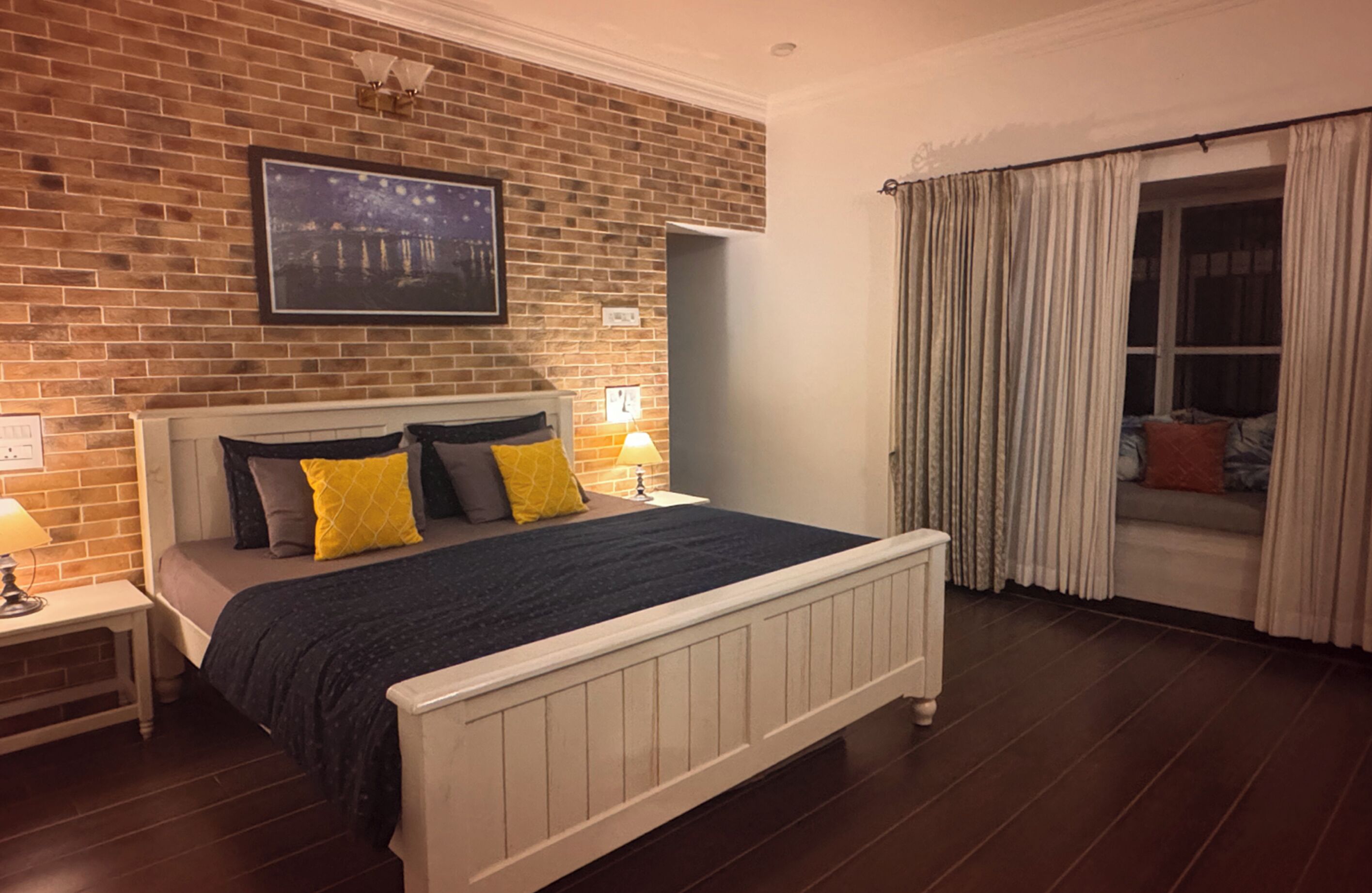 Une chambre avec un mur en briques, un lit avec une tête de lit blanche, une table de chevet, une fenêtre avec des rideaux et un tableau au-dessus du lit.