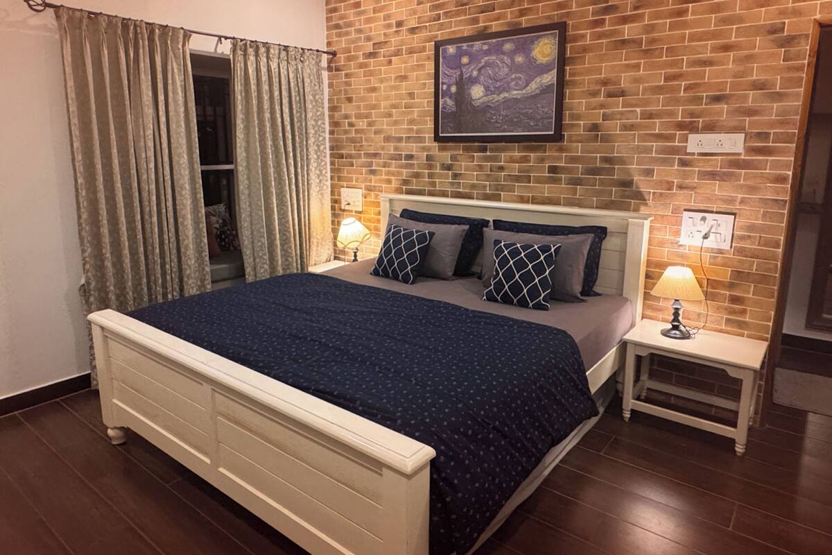 A bedroom with a brick wall, a bed with a dark blue bedspread, two bedside tables with lamps, and a painting above the bed.