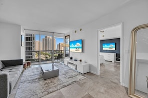 A modern living room with a sofa, coffee table, and TV, large windows with a city view, and a kitchen area.