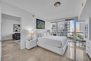 Ein modernes Schlafzimmer mit einem groĂen Bett, einem Nachttisch, einer Lampe, einem GemĂ€lde und einem Balkon mit Blick auf GebĂ€ude.