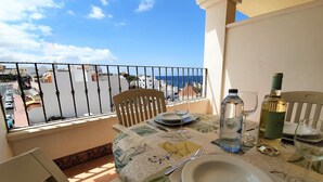 Un balcon avec une table dressée pour deux personnes, donnant sur une zone côtière avec des bâtiments et une vue sur la mer.