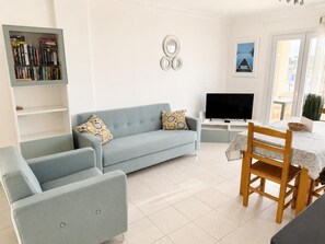 A living room with a sofa, armchair, TV, bookshelf, and dining area with a table and chairs.